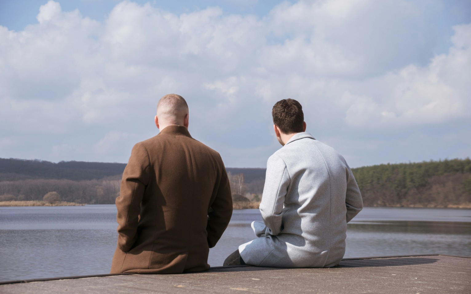 Two men sitting together on a jetty looking out over a lake